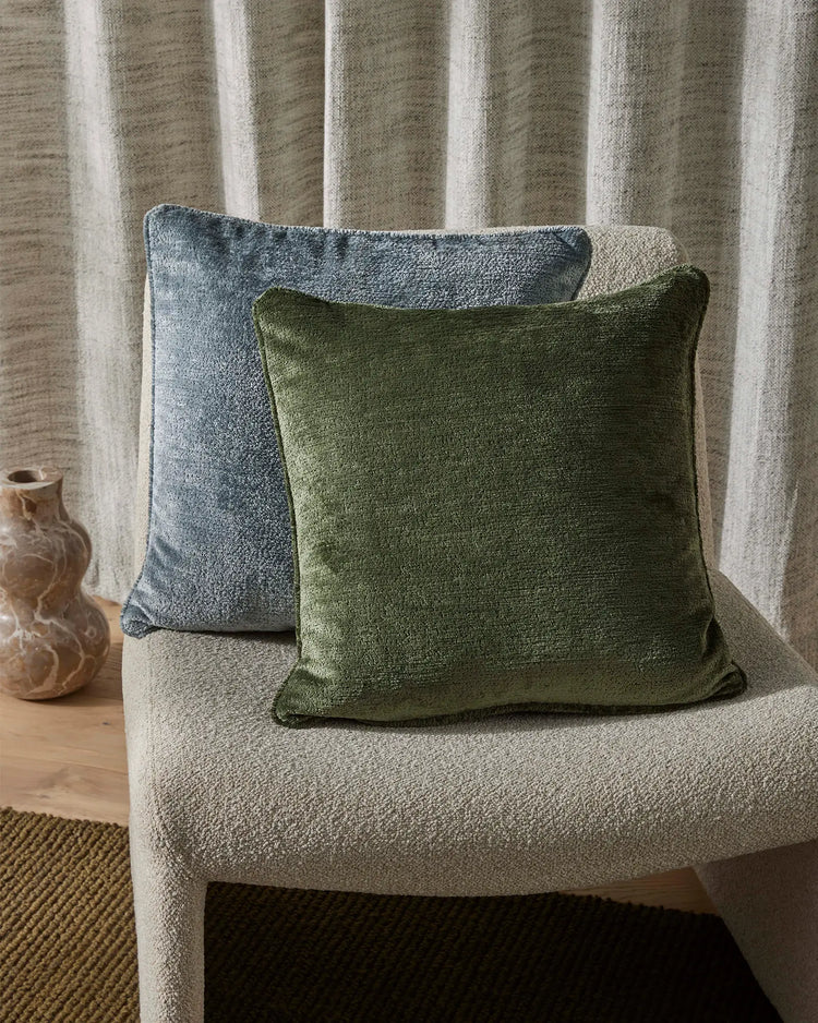 Sage green and slate blue Tempo cushions layered on a natural linen chair with vertical striping, warm timber accents, and textured jute flooring in earthy minimalist styling.