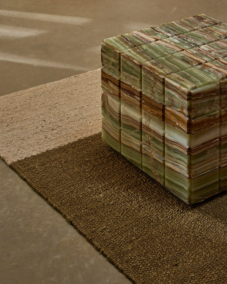 Neutral jute rug in warm taupe tones layered with striped earth-toned textile, creating earthy minimalist aesthetic in concrete space