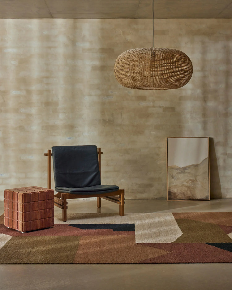 Earthy minimalist interior with geometric terracotta and neutral rug, timber lounge chair, woven pendant and brick-textured wall backdrop