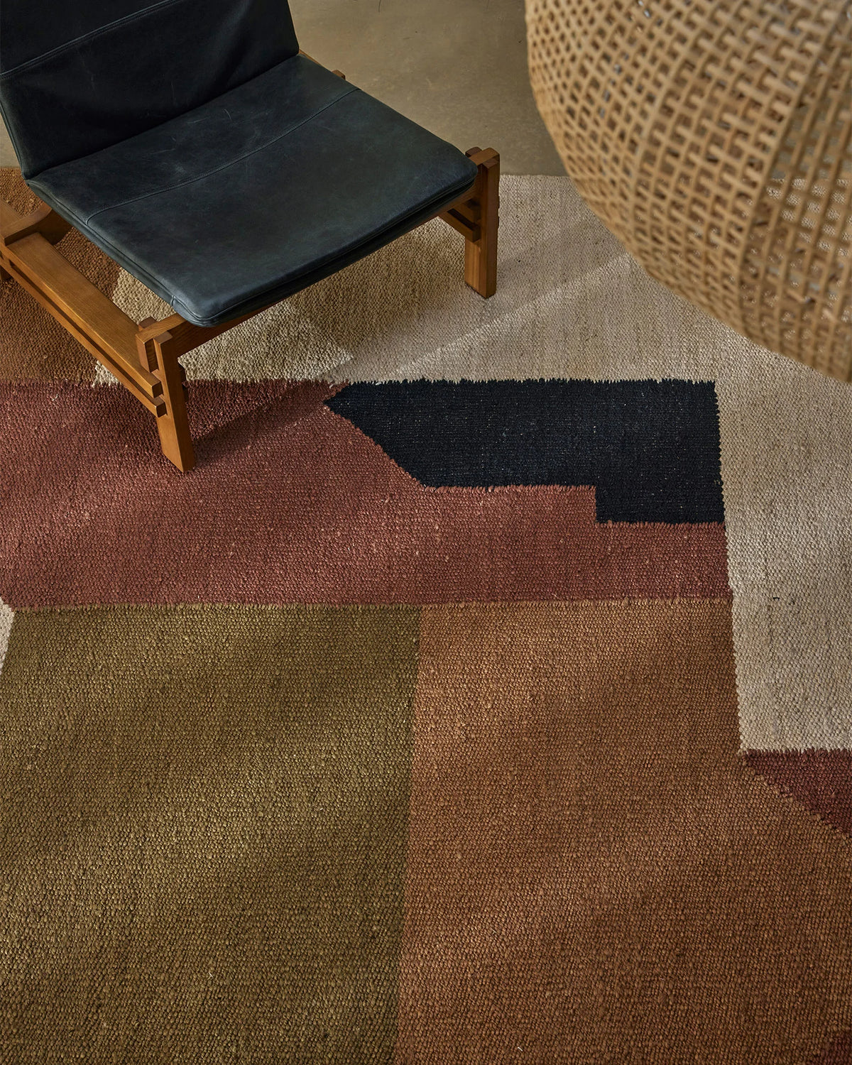 Earthy-toned rug in terracotta, olive and charcoal with geometric colour-block pattern, styled in modern living room with mid-century wooden chair and woven pouf accent