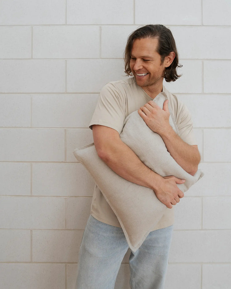 Como Linen cushion in off-white European linen, 50x50cm, held by man against white brick wall by Weave Home