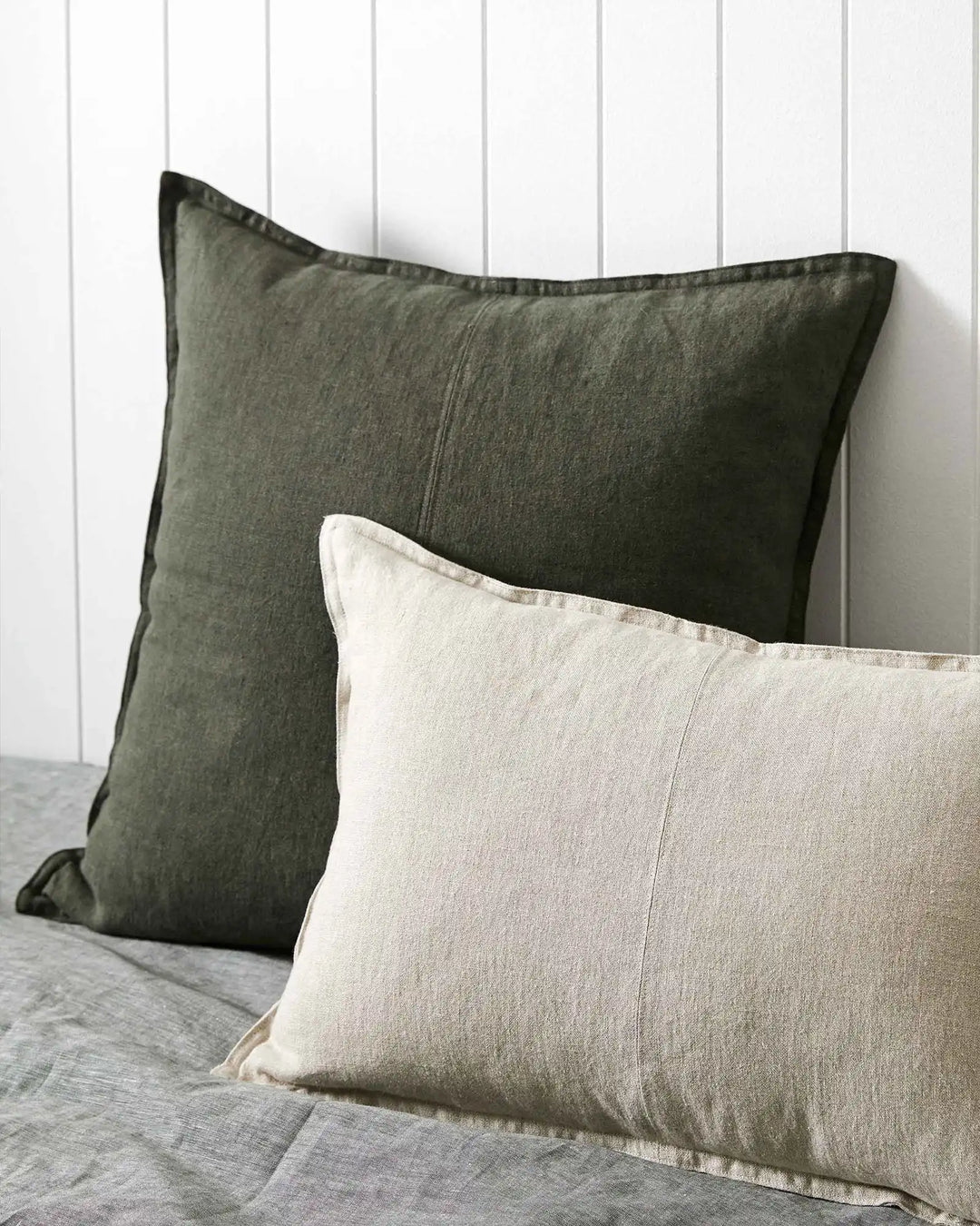Sage and cream linen cushions layered on neutral bedding against white shiplap wall, showcasing relaxed texture and earthy minimalist bedroom styling