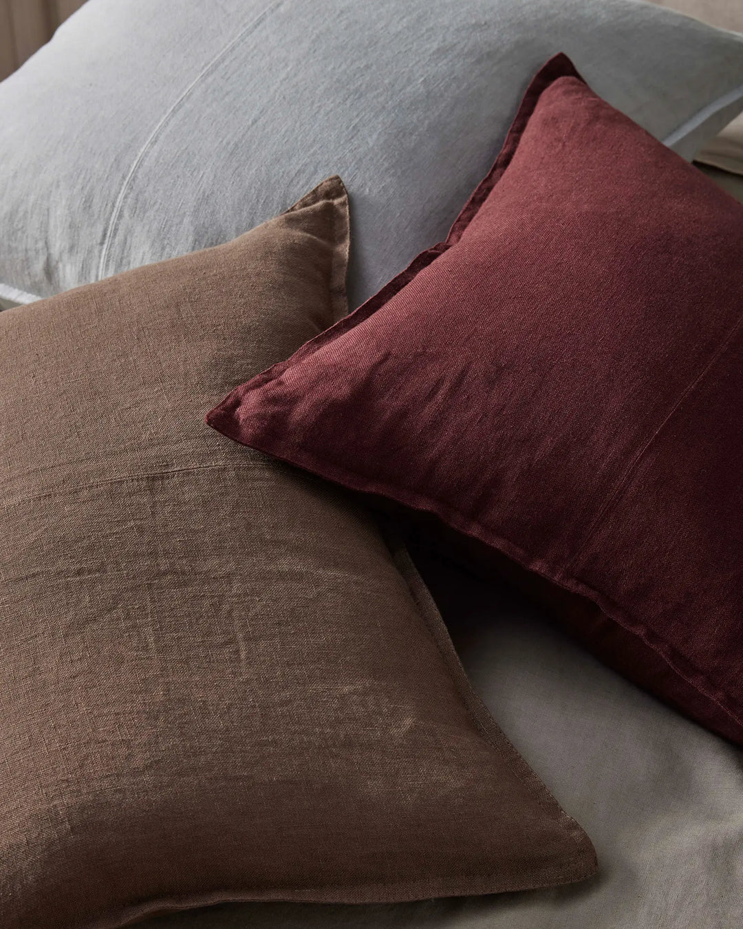 Layered linen cushions in warm mocha and deep garnet tones on a pale grey upholstered chair, creating an earthy, textural palette with natural linen weave detail