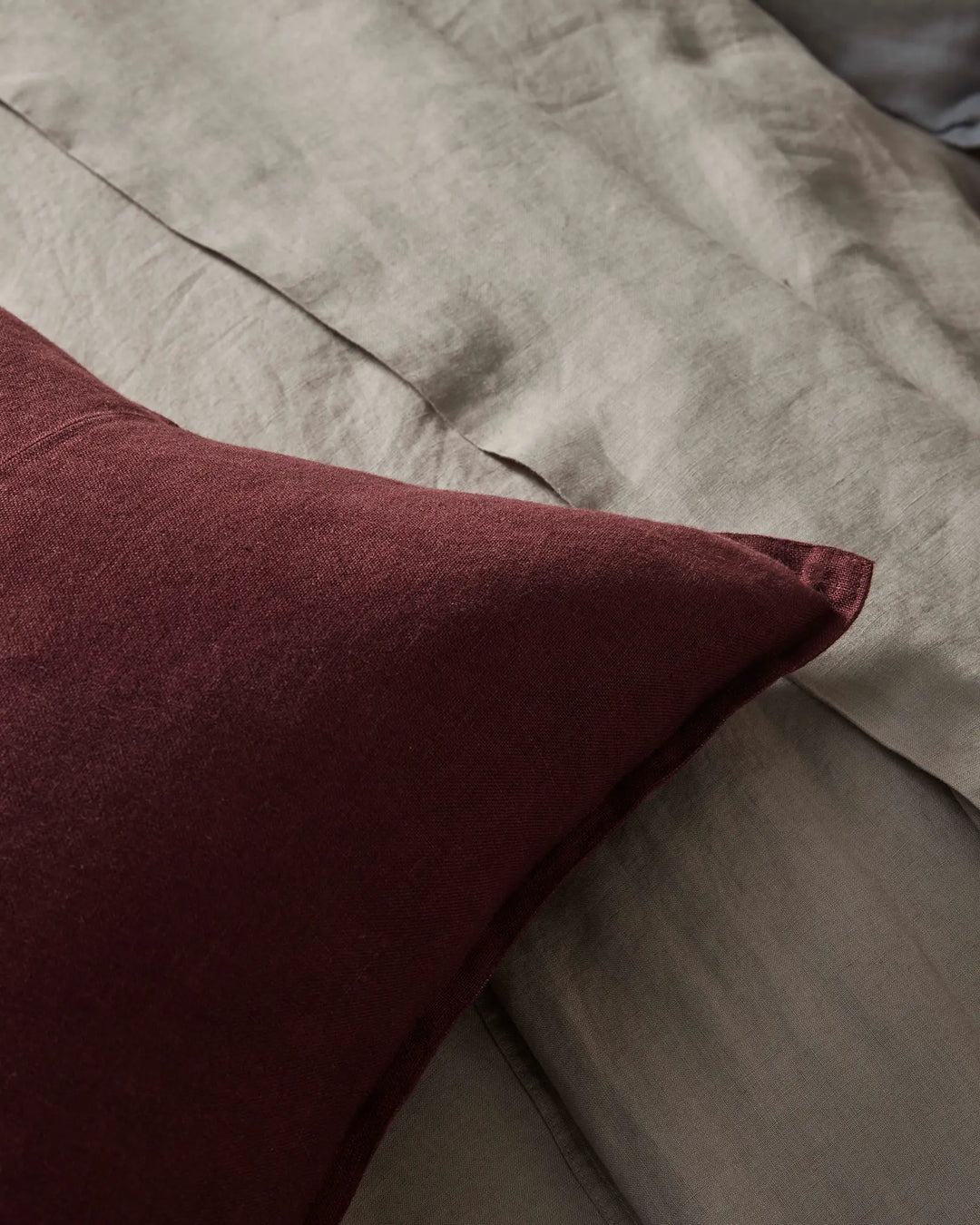 Burgundy linen cushion layered with neutral linen bedding in warm taupe, close-up of textured fabric weave and soft folds for modern bedroom styling