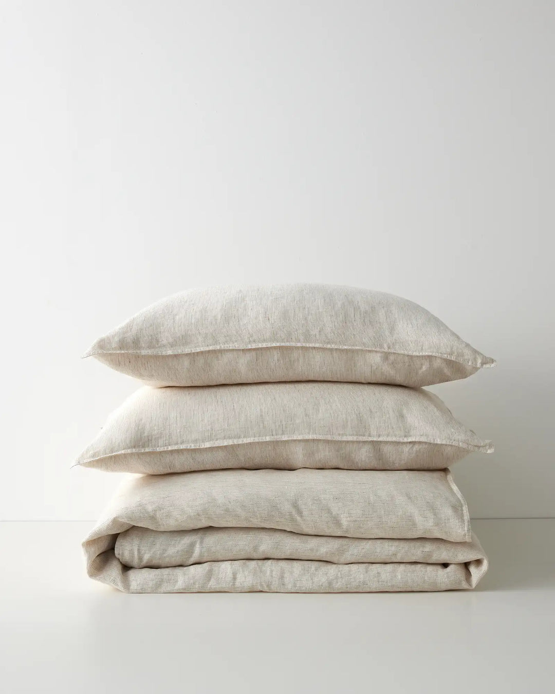 Stacked neutral linen pillows and folded quilt cover in cream and beige tones, showcasing soft texture and minimalist bedroom styling against a light backdrop