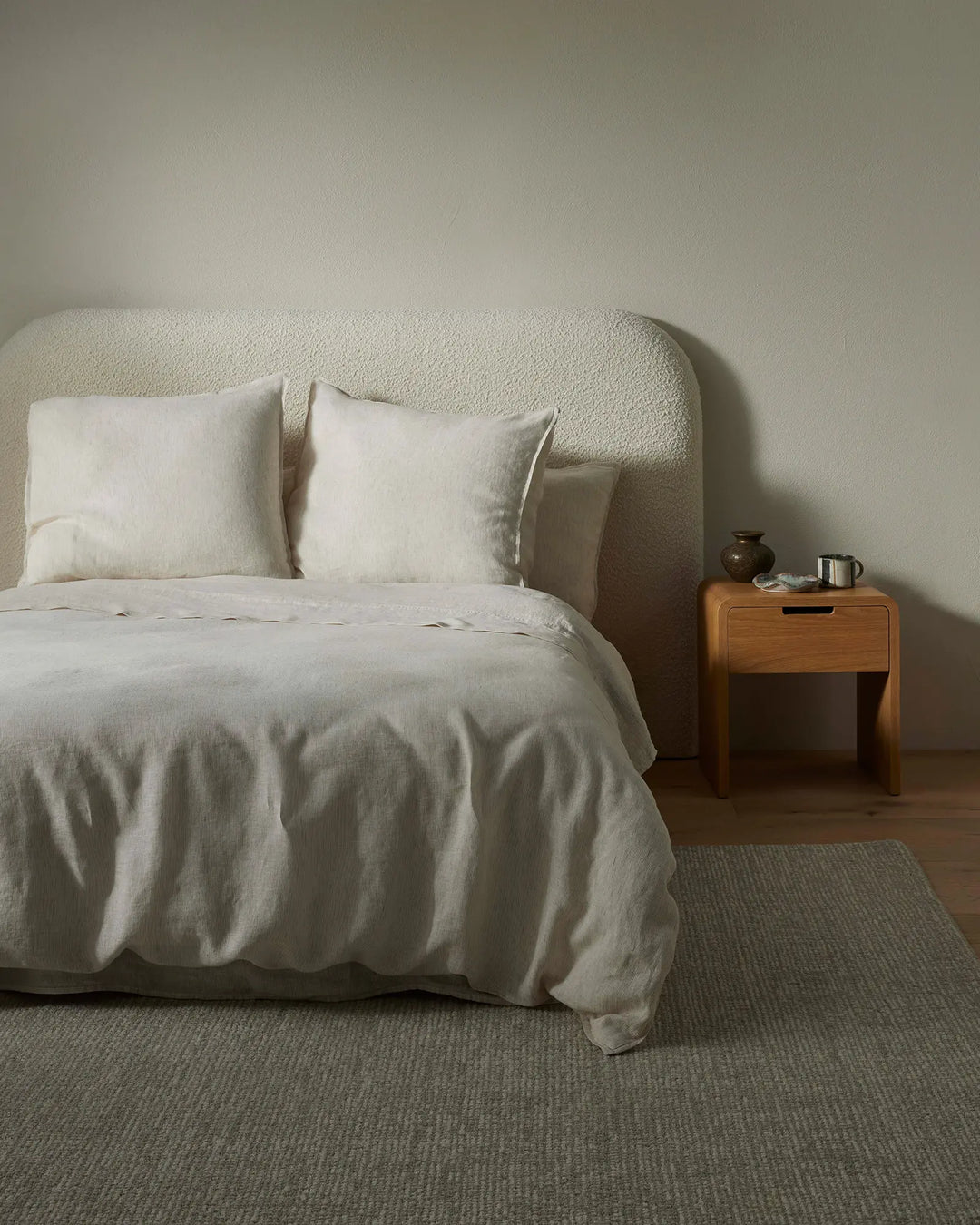Minimalist bedroom with Sova Quilt Cover in soft natural tones, layered with cream linen pillows on a curved upholstered headboard, warm timber bedside table, and neutral textured area rug