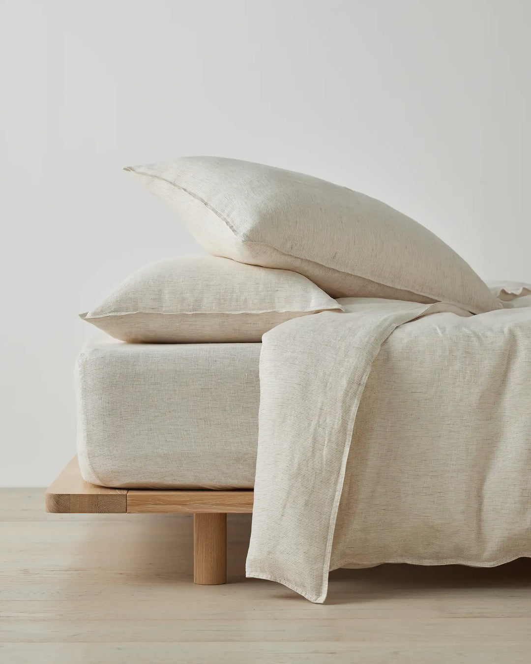 Minimal bedding arrangement with warm cream linen pillows and flat sheet layered over natural wool duvet on blonde timber platform bed against soft grey backdrop