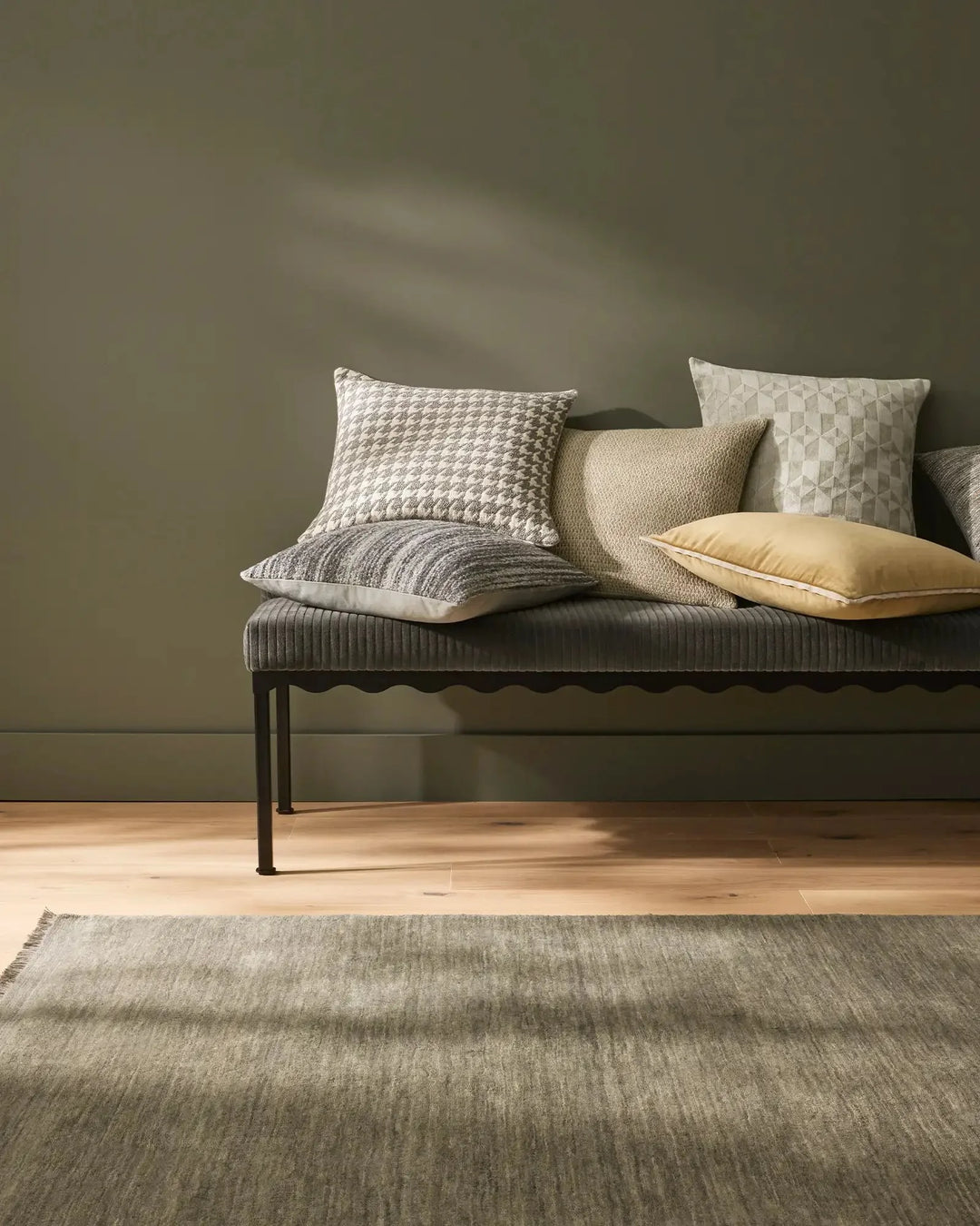 Sage green bedroom styled with textured throw cushions in cream, grey and pale yellow on a ribbed bench, warm timber flooring and neutral area rug creating calm, minimalist aesthetic