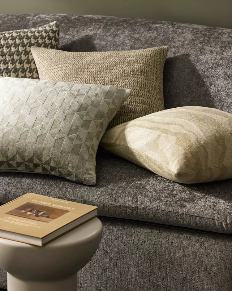 Neutral living room styled with layered cushions in cream, natural, and charcoal tones; textured linens and viscose blends arranged on a grey upholstered sofa with a book nearby