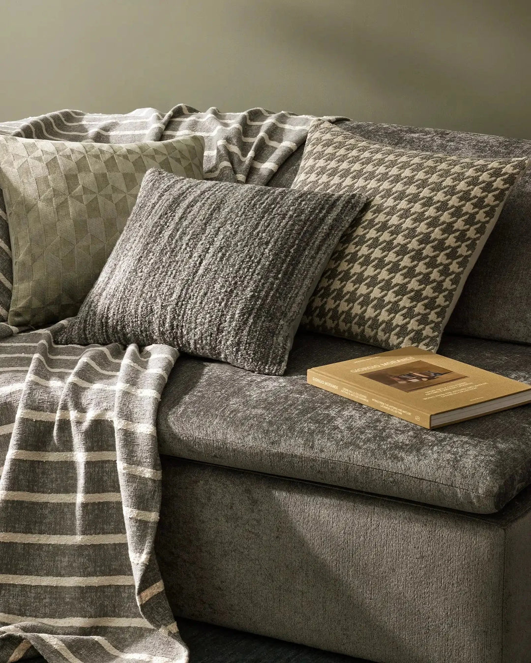 Neutral textured cushions and Piazza throw layered on charcoal sofa, earthy palette of sage, taupe and cream with geometric and knit textures, minimalist styling with gold-bound book