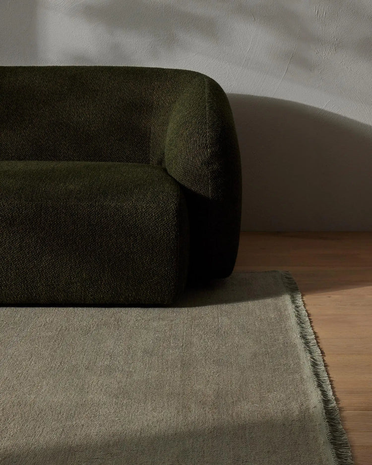 Sage green armchair with rolled arm beside a natural linen rug in minimalist bedroom setting, highlighting textural woven fabrics and warm timber floor
