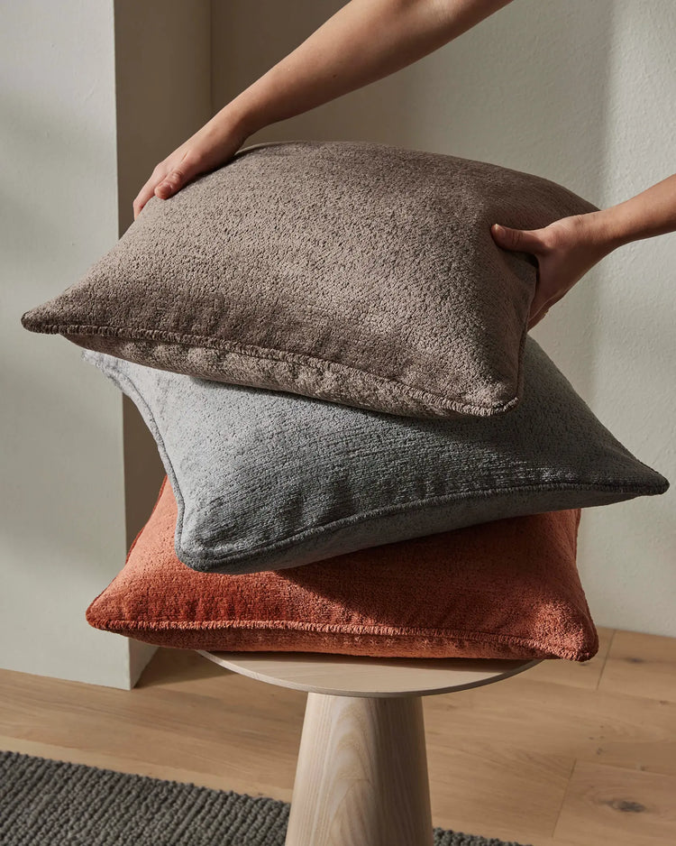 Stacked Tempo cushions in cocoa, spice, and steel tones styled on wooden side table, showcasing textured weave fabrics in warm earth and cool grey palette