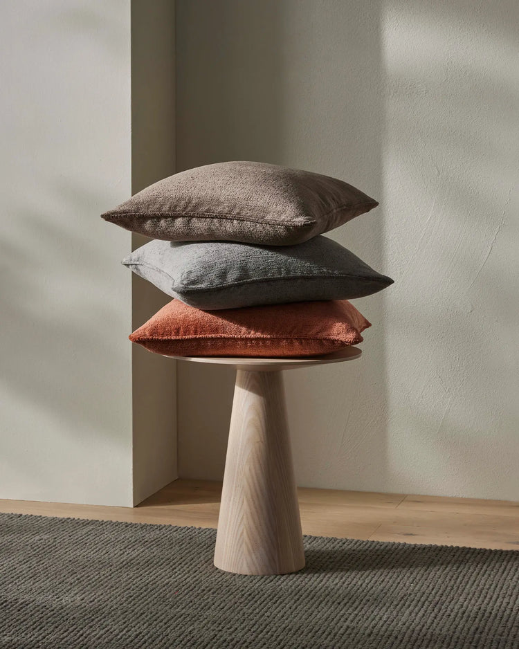 Stacked cushions in warm spice, cool steel grey and cocoa tones on natural wood pedestal table, minimalist neutral interior styling