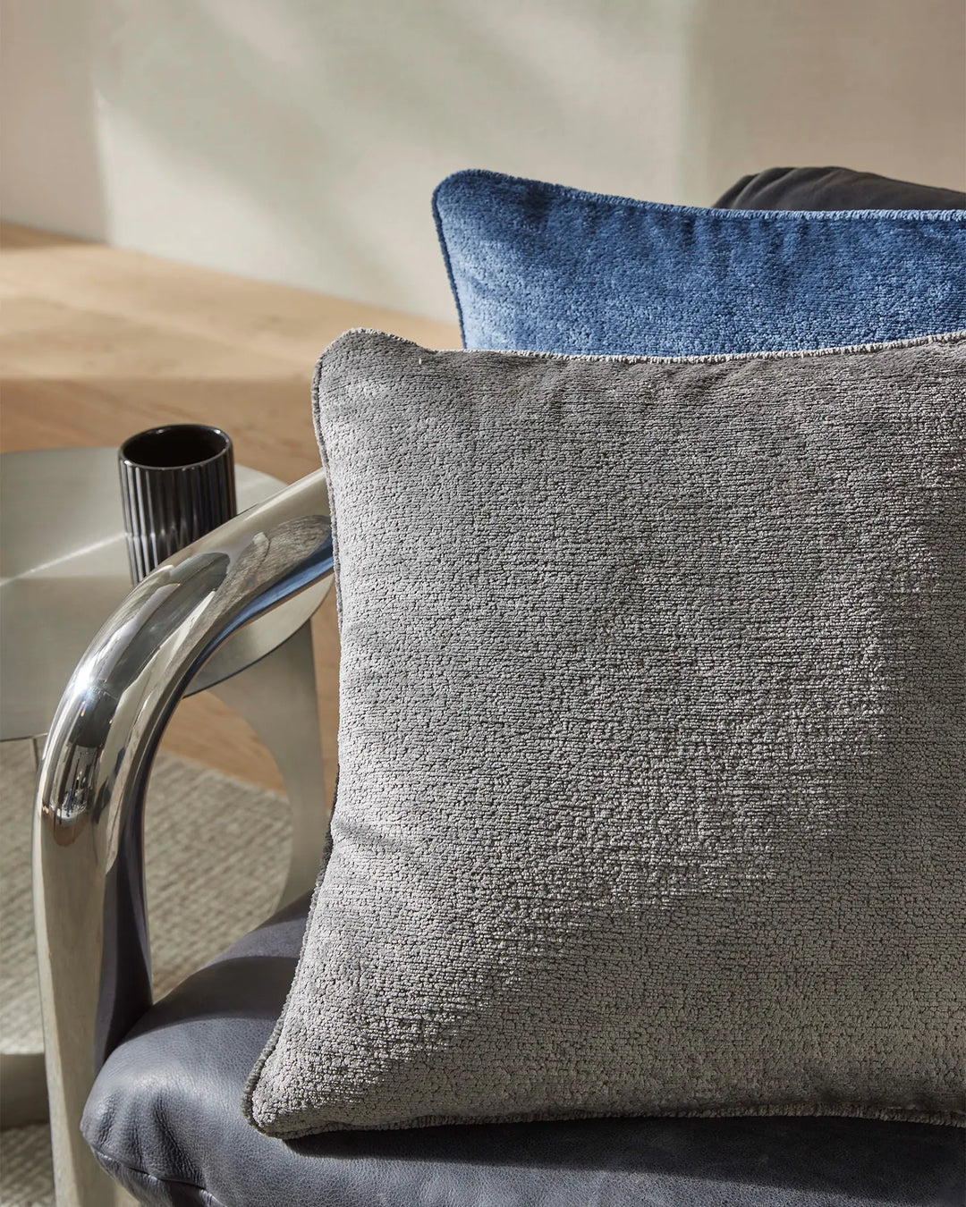 Neutral and blue textured cushions layered on a modern chair with chrome details, creating a sophisticated minimalist aesthetic with warm timber and soft natural light