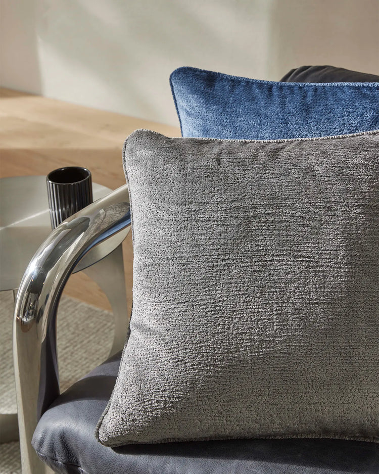 Neutral and blue textured cushions layered on a modern chair with chrome details, creating a sophisticated minimalist aesthetic with warm timber and soft natural light