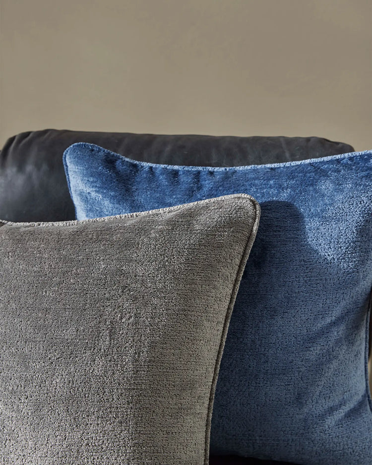 Layered Tempo cushions in steel grey and marine blue on dark sofa, showcasing textured woven fabrics and neutral tones against pale wall backdrop