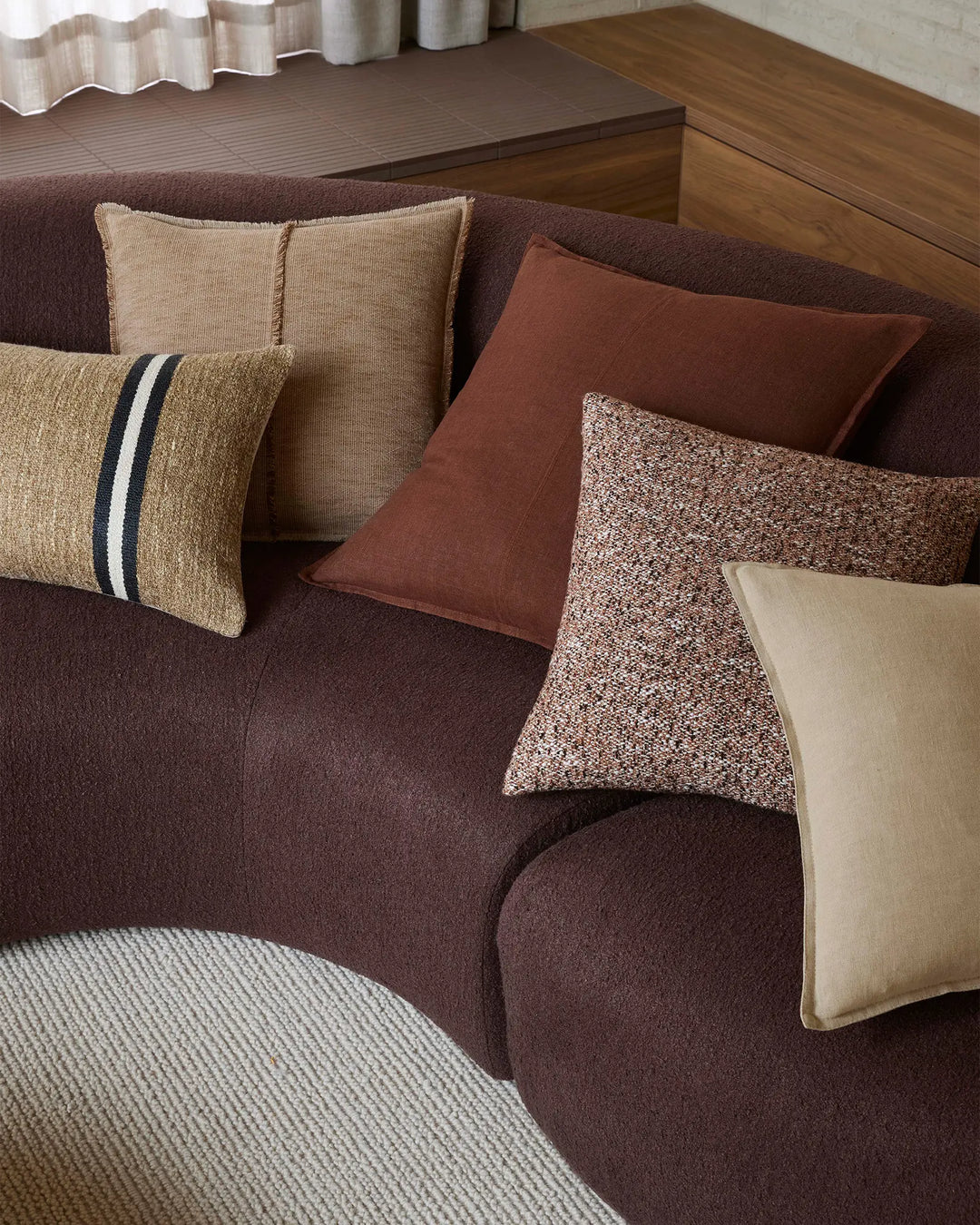 Warm-toned living room styled with earthy cushions in chocolate, terracotta and natural linen textures, layered on a deep brown sofa with wood furnishings and soft neutral accents