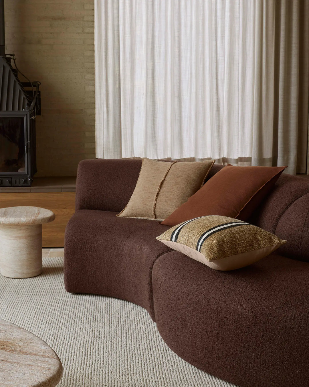 Warm earthy living room with chocolate brown curved sofa layered with terracotta, natural linen, and gold striped cushions, cream textured rug, and soft white striped curtains filtering light
