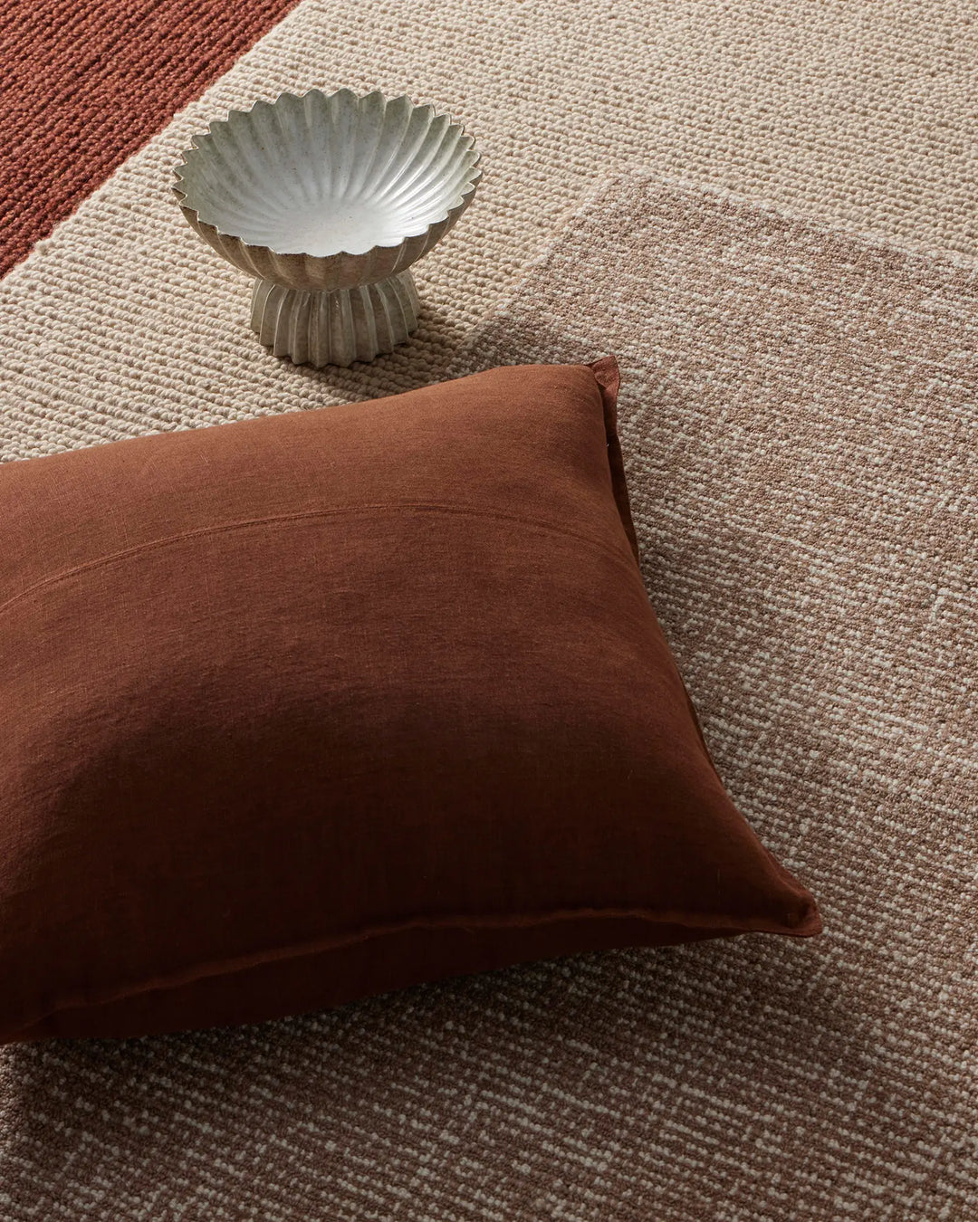 Rust and cream linen cushion and woven rugs layered on natural textiles, with sculptural ceramic bowl, earthy tonal palette styling