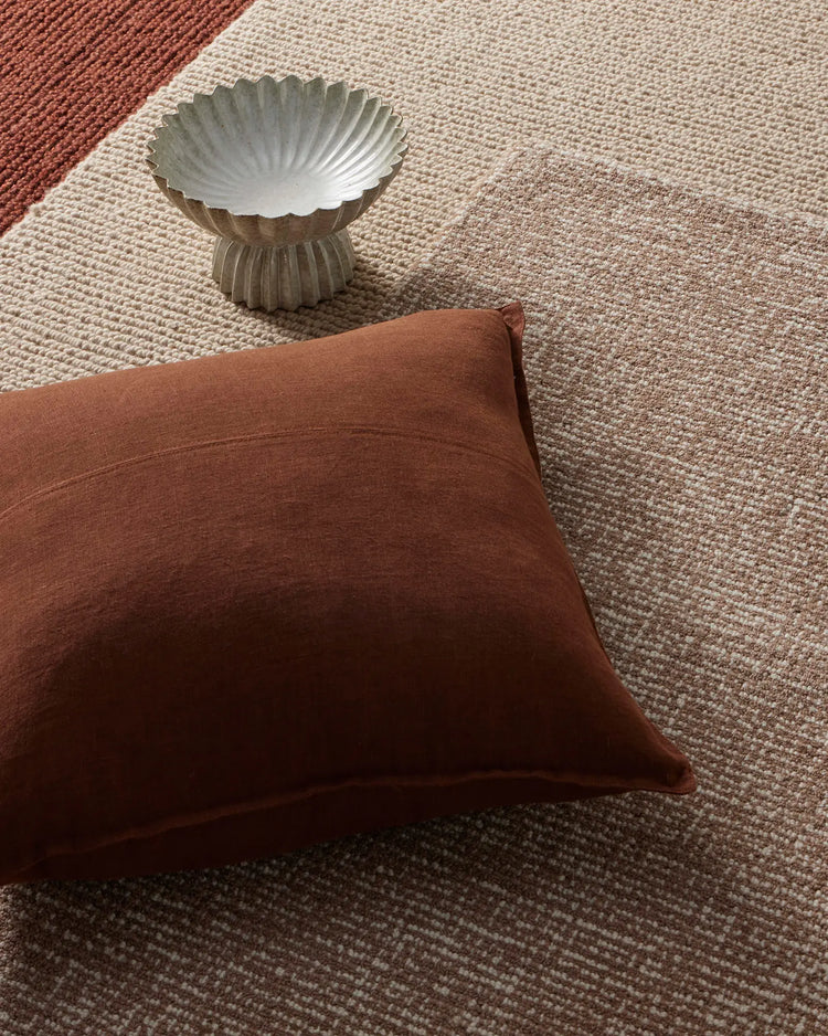Rust and cream linen cushion and woven rugs layered on natural textiles, with sculptural ceramic bowl, earthy tonal palette styling