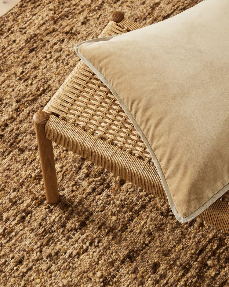 Neutral linen cushion on woven chair with natural wool rug, earthy tones and textured layering in organic minimalist interior styling