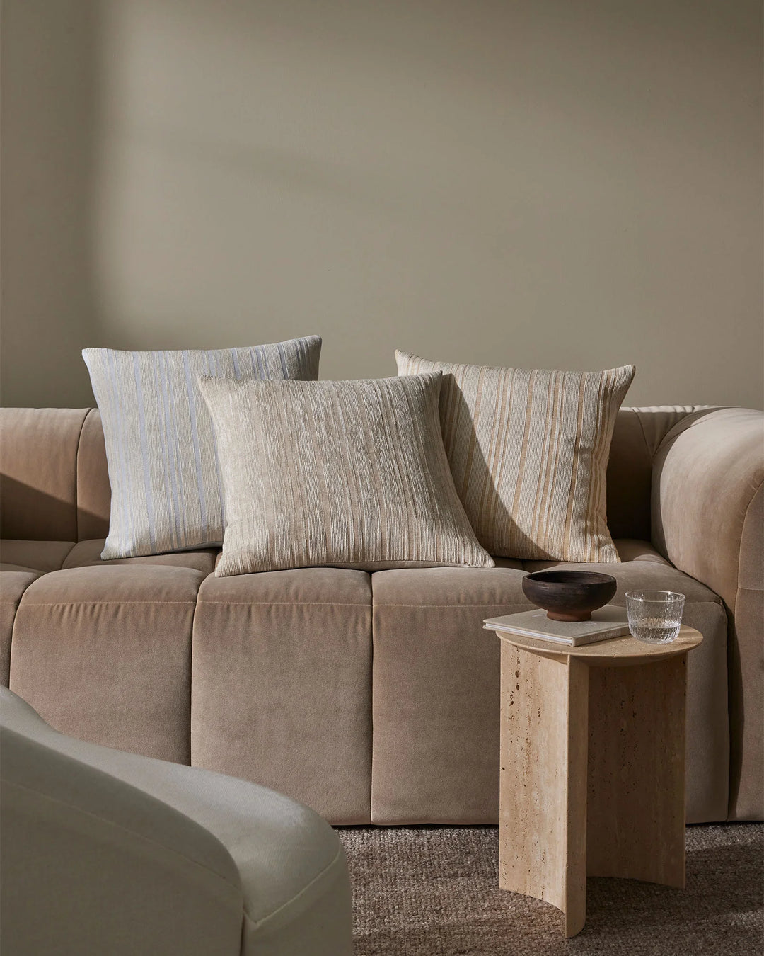 Neutral living room with beige curved sofa styled with three textured linen cushions in glacier, butter and natural tones, travertine side table, warm earthy palette