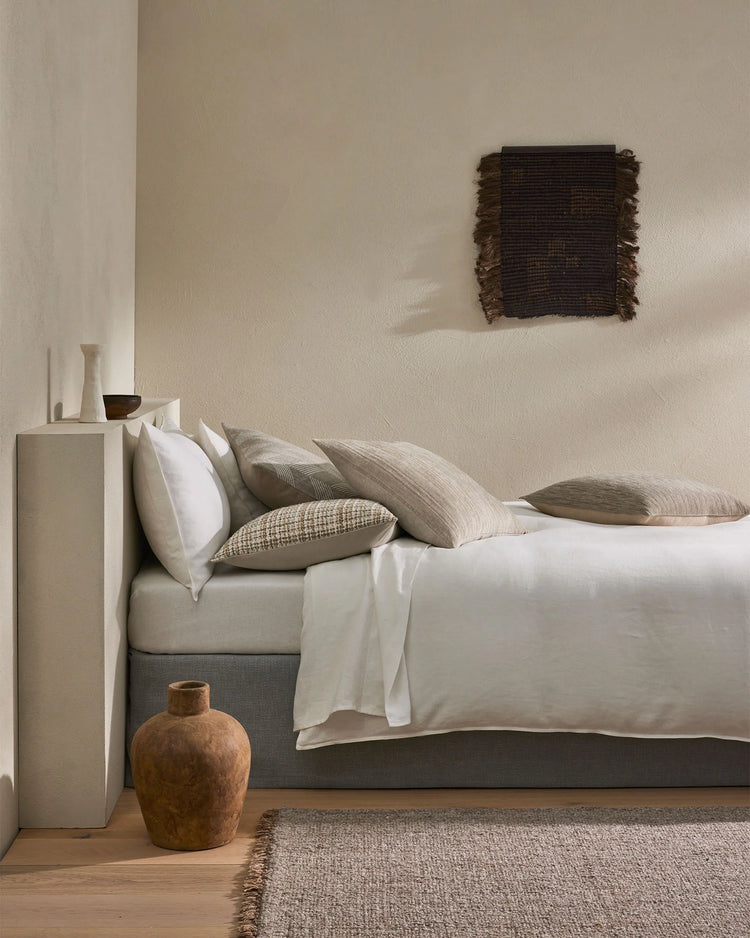 Neutral minimalist bedroom with layered cushions in cream, beige and natural tones, textured wall art, terracotta vessel and linen bedding on platform bed