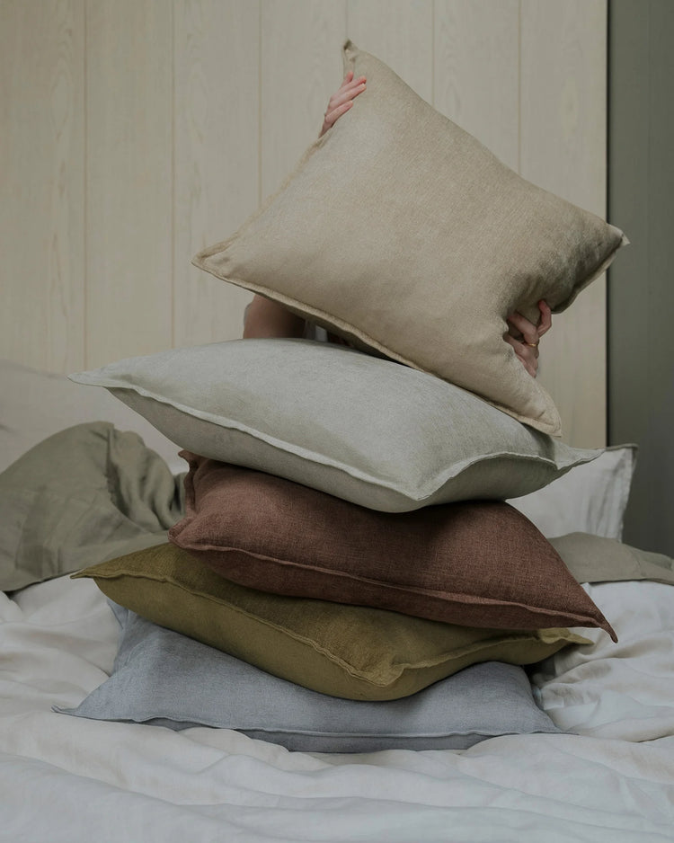 Stacked Fiore cushions in neutral earth tones—sage, toffee, ecru, and breeze—showcase linen viscose blend textures against white panelled walls and bedding