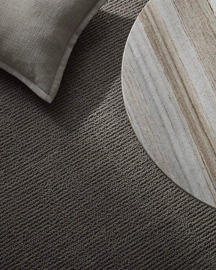 Charcoal wool rug with natural texture and weave detail, styled with grey linen cushion and whitewashed round side table in minimalist Scandinavian interior setting