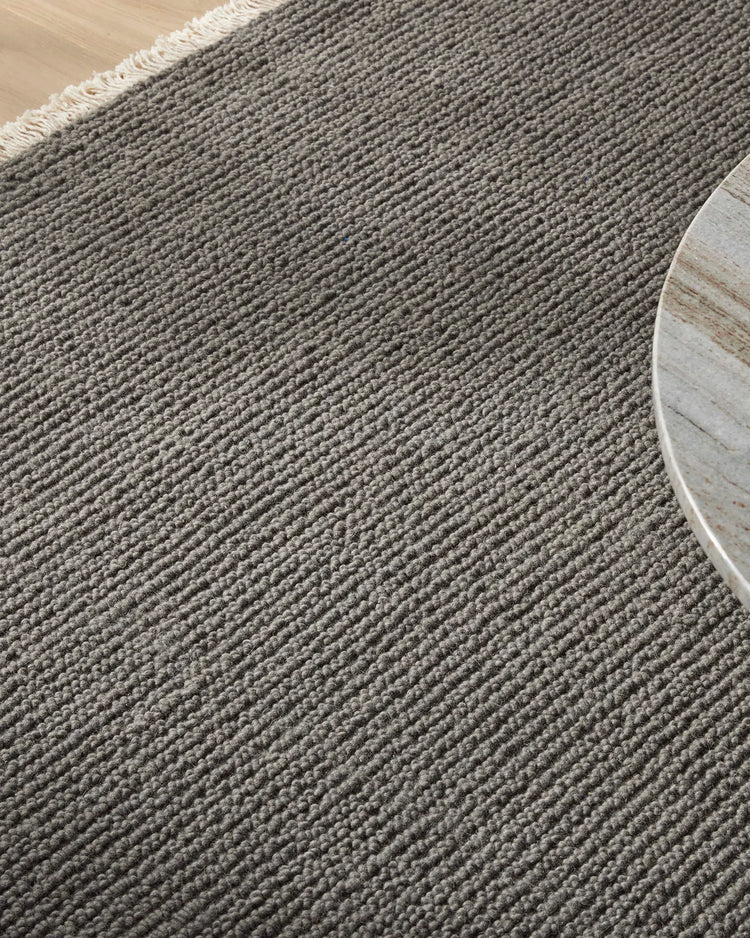 Grey wool rug with chunky woven texture and cream fringe edge on light timber floor beside white round side table