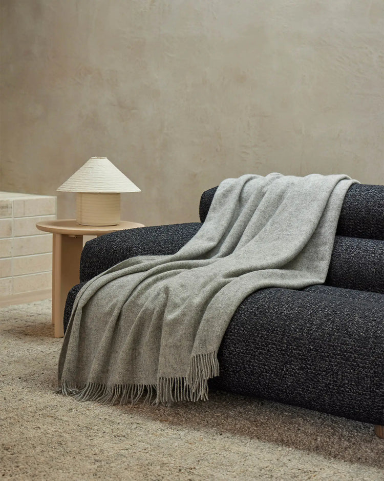 Light grey wool throw draped over charcoal sofa with natural wood side table and geometric lamp in warm minimalist living room setting