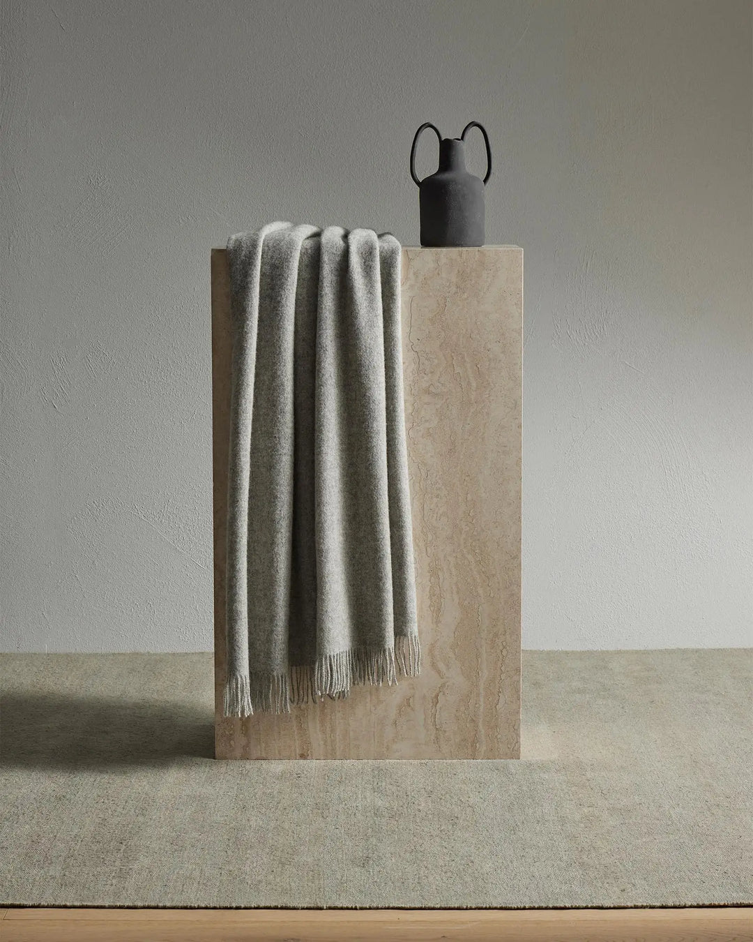 Neutral minimalist styling with draped grey wool throw over travertine block, black ceramic vessel, and soft stone textures against white wall