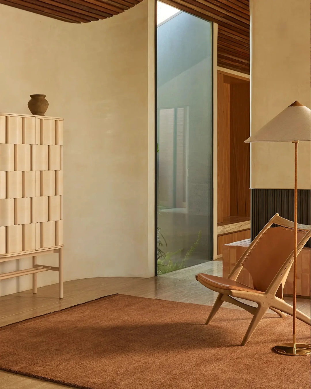 Warm terracotta rug anchoring a minimalist mid-century living space with cream walls, wooden furnishings, and soft overhead lighting; Silvio in Sienna.